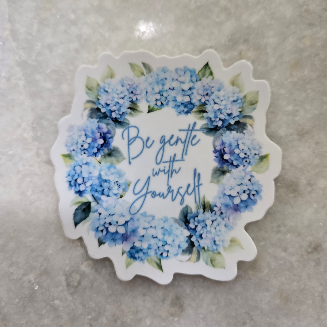 A scalloped die-cut sticker featuring a watercolor floral wreath in shades of blue and purple with green leaves, surrounding the cursive text 'Be gentle with Yourself' on a white background, placed on a light gray marble surface. Pink Salt Riot