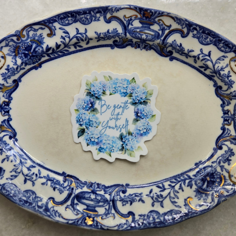 An oval ceramic plate with a blue and gold floral scroll pattern, featuring a central sticker that reads 'Be sure to carry on'. The plate is photographed on a light gray marble surface. Pink Salt Riot