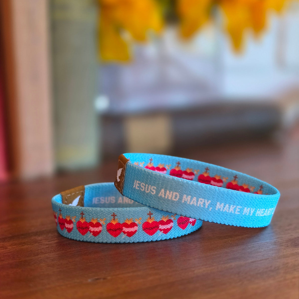 Two light blue woven wristbands with red and white Sacred Heart motifs and white text, resting on a wooden tabletop- Pink Salt Riot 