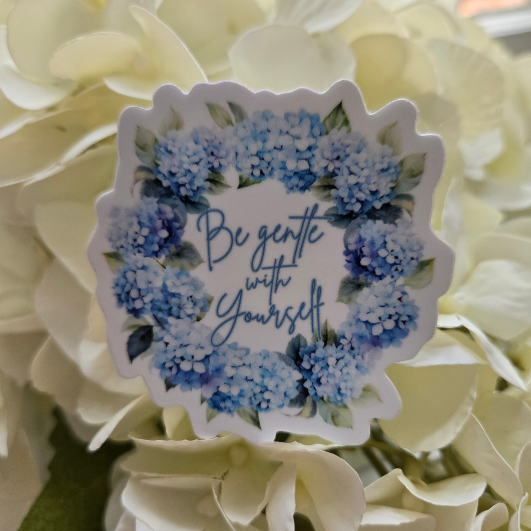 A scalloped die-cut sticker featuring the phrase 'Be gentle with Yourself' in blue cursive, surrounded by a watercolor-style floral wreath of blue hydrangeas and green leaves, shown resting on cream-colored hydrangea petals. Pink Salt Riot